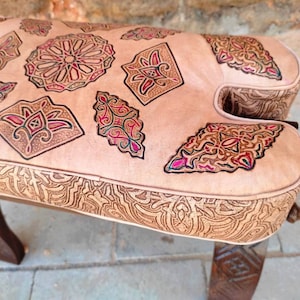 May include: A light brown leather ottoman with intricate carved designs in shades of red, green, and brown. The ottoman has a wooden frame with carved details.