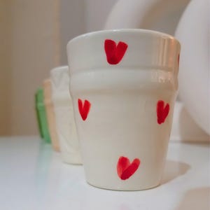 Peut inclure: Une tasse en céramique blanche avec des motifs de cœurs rouges. La tasse est de forme simple, cylindrique avec un fond arrondi.