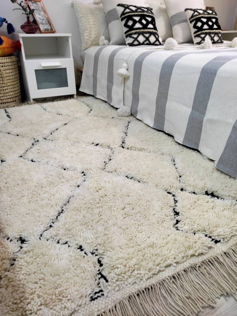 May include: A white shag rug with a black diamond pattern. The rug has a fringe border and is laid out on a hardwood floor.