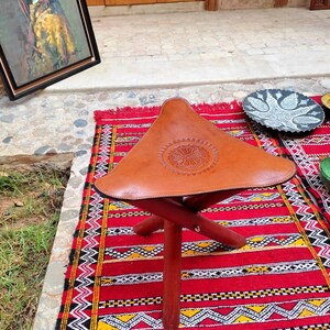 May include: A brown leather folding stool with a decorative design on the seat. The stool is sitting on a red, black, and white patterned rug.