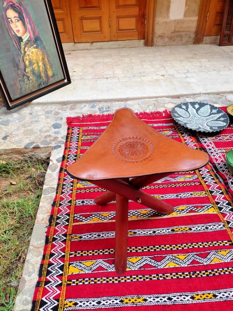 May include: A brown leather folding stool with a decorative design on the seat. The stool is sitting on a red, black, and white patterned rug.