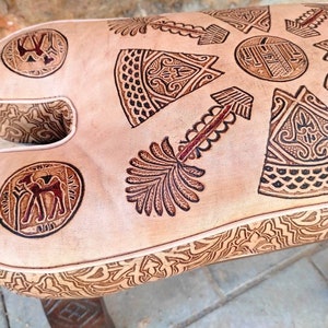 May include: A light brown leather ottoman with intricate geometric patterns embossed on the top. The ottoman has a rounded, saddle-like shape and is supported by wooden legs.