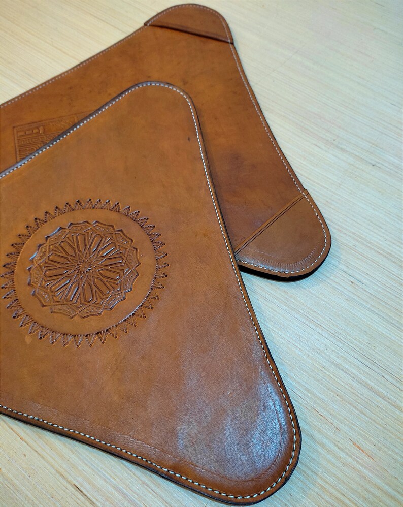 May include: Two brown leather triangular coasters with decorative stitching around the edges. One coaster features a circular design with intricate details.