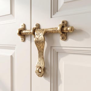 Peut inclure: Un loquet de porte en laiton orné d'un motif de feuilles gravé sur la barre horizontale et la poignée verticale. La quincaillerie est fixée par quatre vis apparentes sur une porte blanche à panneaux, assurant un mécanisme de fermeture.