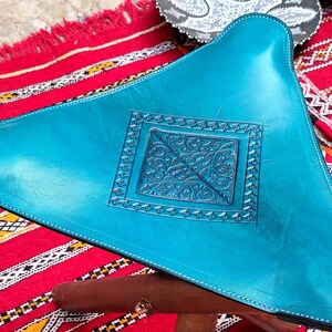 Op de afbeelding: Een turquoise leren poef met een decoratief reli&euml;f in het midden. De poef staat op een rood-wit gestreept tapijt.