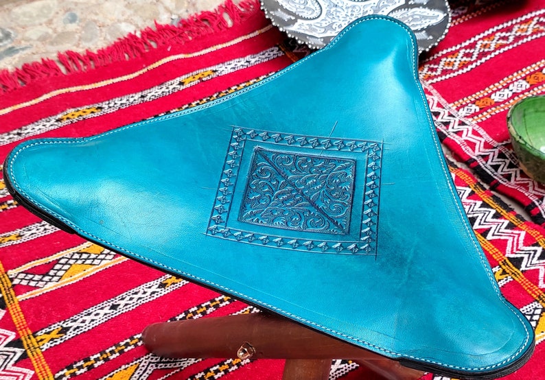 Op de afbeelding: Een turquoise leren poef met een decoratief reli&euml;f in het midden. De poef staat op een rood-wit gestreept tapijt.