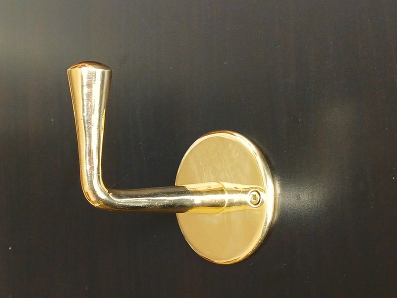 May include: A gold-toned metal hook with a rounded handle and a circular base. The hook is mounted on a dark surface.