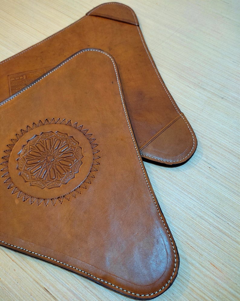 Op de afbeelding: Twee bruine, driehoekige leren onderzetters met decoratieve stiksels rond de randen. Een onderzetter heeft een cirkelvormig ontwerp met ingewikkelde details.
