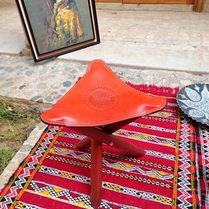 Op de afbeelding: Een rode leren vouwbare kruk met een decoratief ontwerp op de zitting. De kruk staat op een rood en zwart gestreept tapijt.