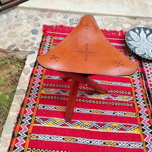 May include: A brown leather three-legged stool with a triangular seat and decorative stitching. The stool is sitting on a red, white, and black patterned rug.