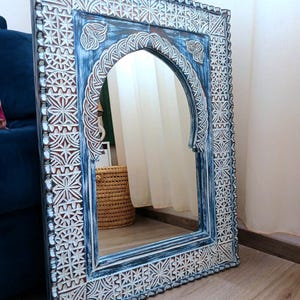 May include: A blue and white carved wooden mirror with an arched top and intricate geometric patterns. The mirror is leaning against a wall.