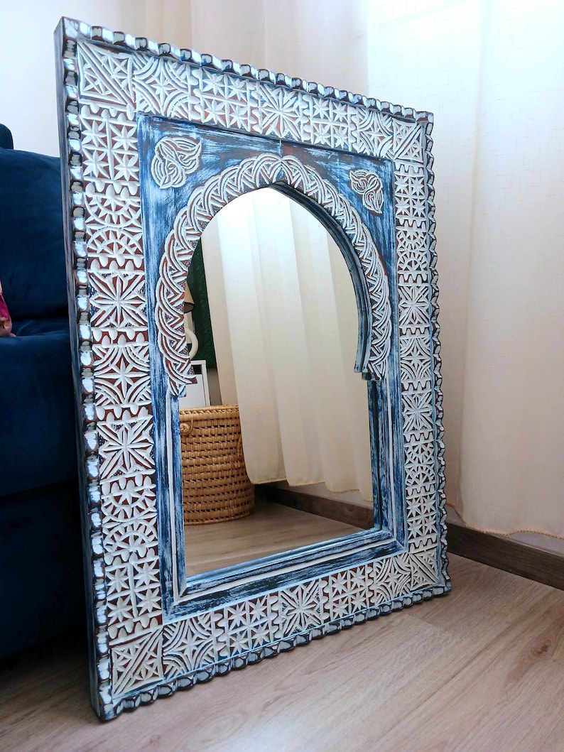 May include: A blue and white carved wooden mirror with an arched top and intricate geometric patterns. The mirror is leaning against a wall.