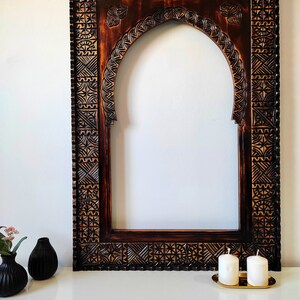 May include: A dark brown wooden frame with intricate carvings, featuring an arched opening. The frame is sitting on a white surface with two white candles on a gold tray.