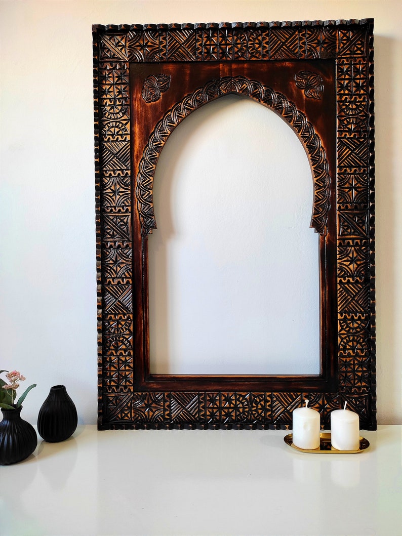May include: A dark brown wooden frame with intricate carvings, featuring an arched opening. The frame is sitting on a white surface with two white candles on a gold tray.