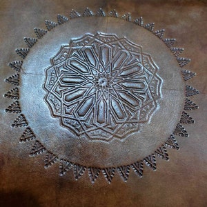 May include: A brown leather cushion with a circular, embossed design in the center. The design features a geometric pattern with a star in the middle.