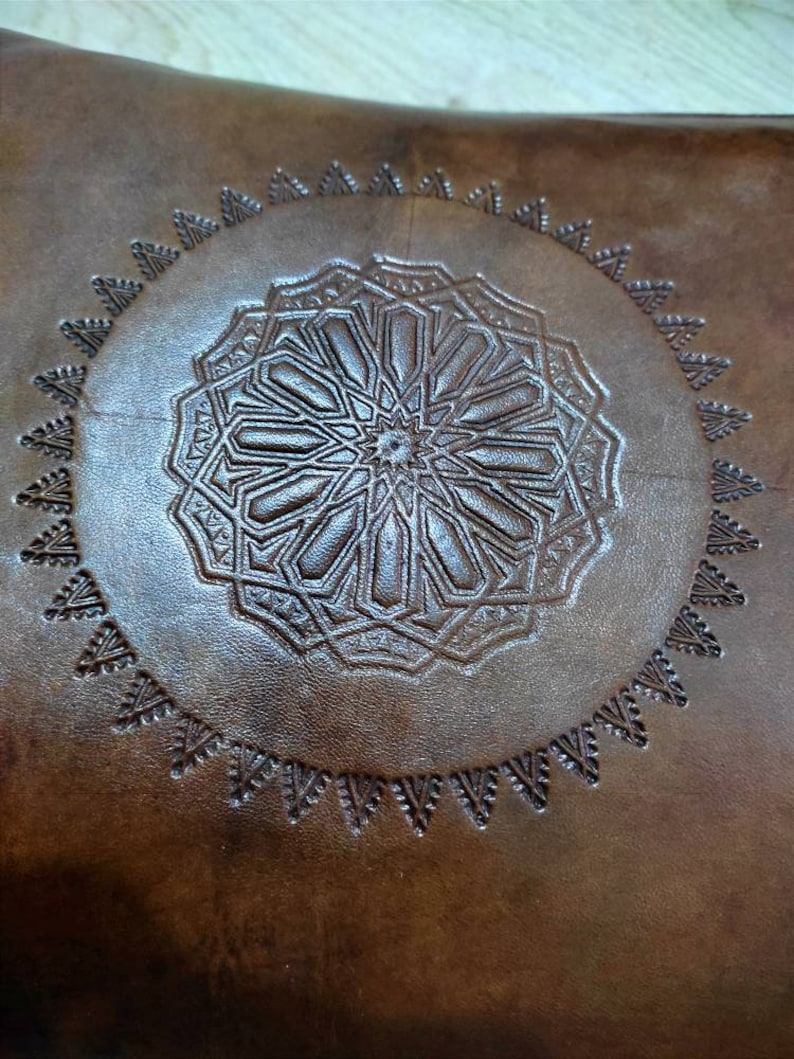 May include: A brown leather cushion with a circular, embossed design in the center. The design features a geometric pattern with a star in the middle.