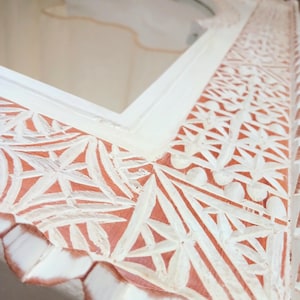 May include: White wooden mirror with intricate carved details. The frame has a distressed white finish with a hint of pink showing through the carvings. The mirror has an arched top.