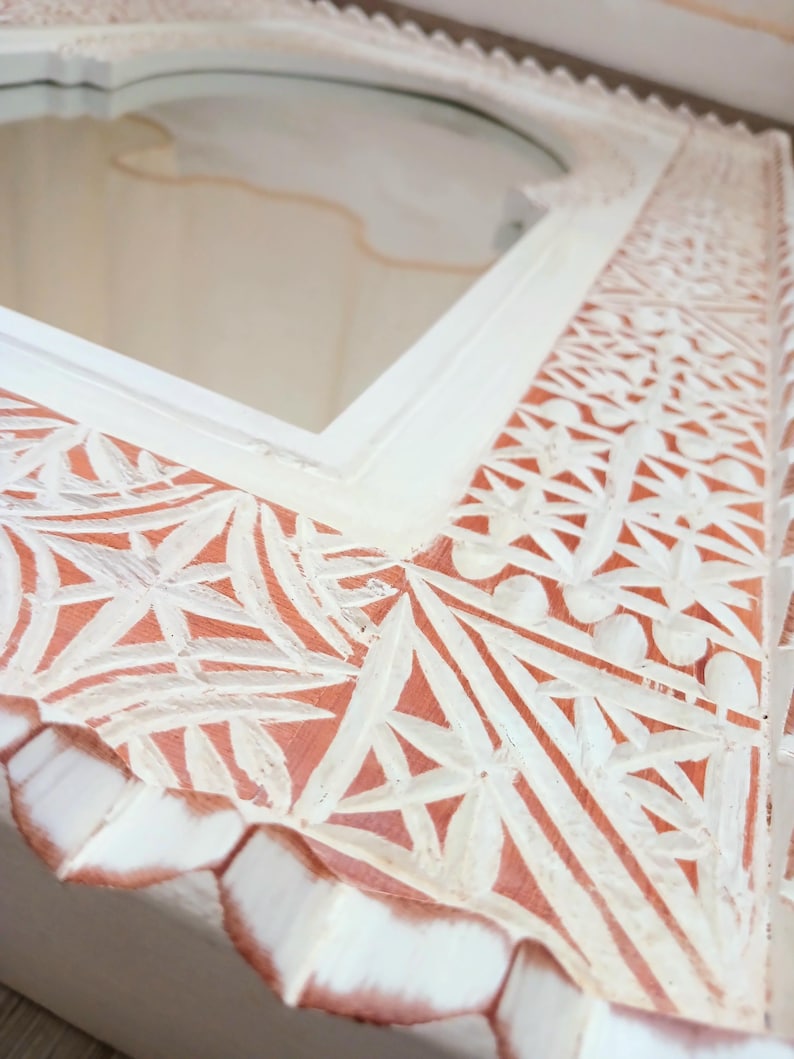 May include: White wooden mirror with intricate carved details. The frame has a distressed white finish with a hint of pink showing through the carvings. The mirror has an arched top.