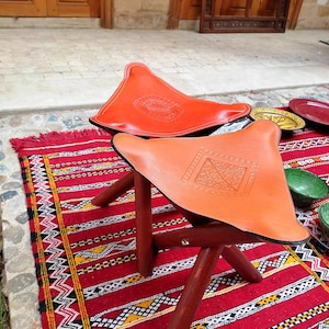Op de afbeelding: Twee oranje Marokkaanse poufs van leer met ingewikkelde ontwerpen op een rood-zwart gepatroneerd tapijt. De poufs zijn gemaakt van leer en hebben een houten frame.