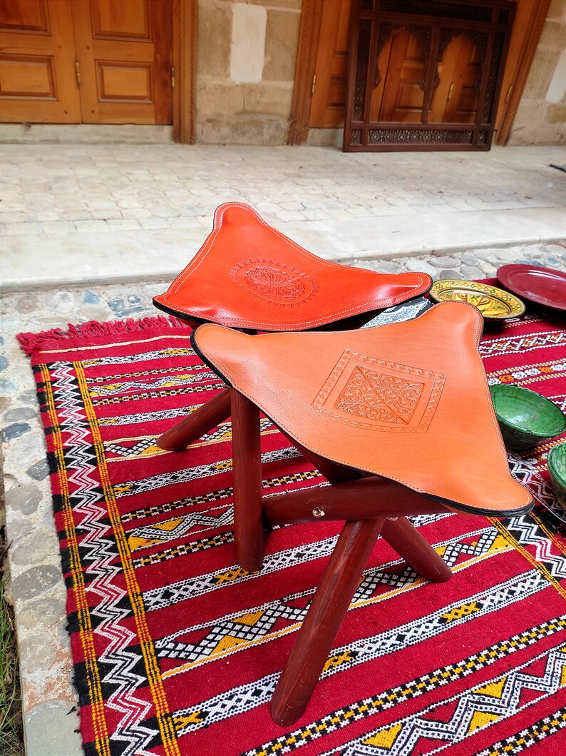 Op de afbeelding: Twee oranje Marokkaanse poufs van leer met ingewikkelde ontwerpen op een rood-zwart gepatroneerd tapijt. De poufs zijn gemaakt van leer en hebben een houten frame.