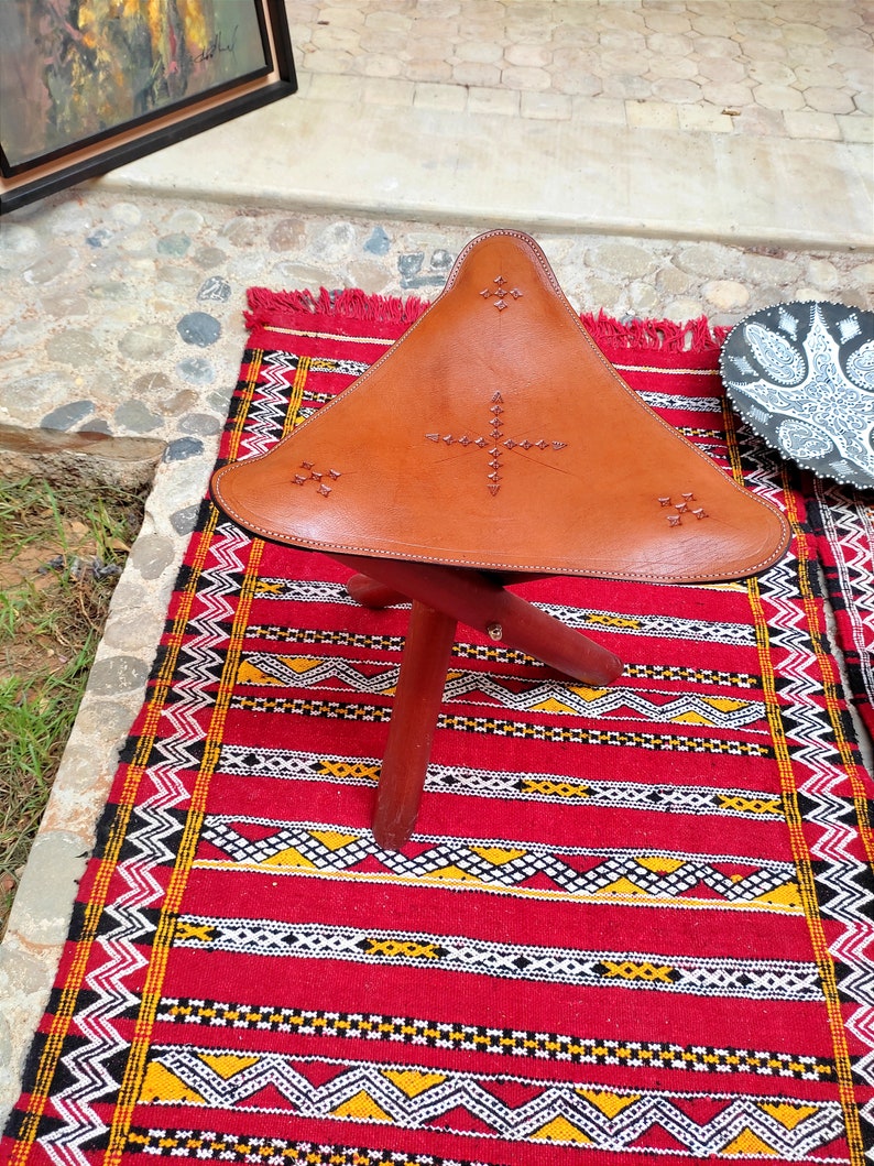 May include: A brown leather triangular stool with decorative stitching and a wooden base. The stool is sitting on a red and yellow patterned rug.