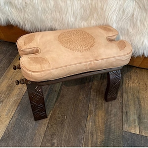 May include: A light brown leather stool with a carved wooden frame. The seat has a circular embossed design in the center.