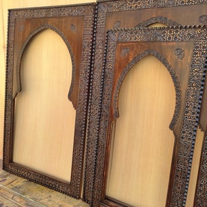 May include: Several dark brown wooden frames with an arched top and intricate carved designs. The frames are empty, with a light tan background visible through the openings. The frames are of varying sizes and are leaning against each other.