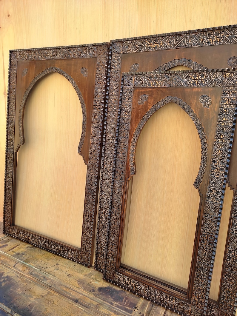 May include: Several dark brown wooden frames with an arched top and intricate carved designs. The frames are empty, with a light tan background visible through the openings. The frames are of varying sizes and are leaning against each other.