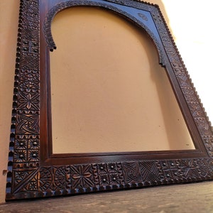 May include: Dark brown wooden frame with intricate carved geometric patterns. The frame has an arched top and a rectangular shape, with detailed carvings along the edges. The wood has a rich, warm tone, and the frame appears to be empty.