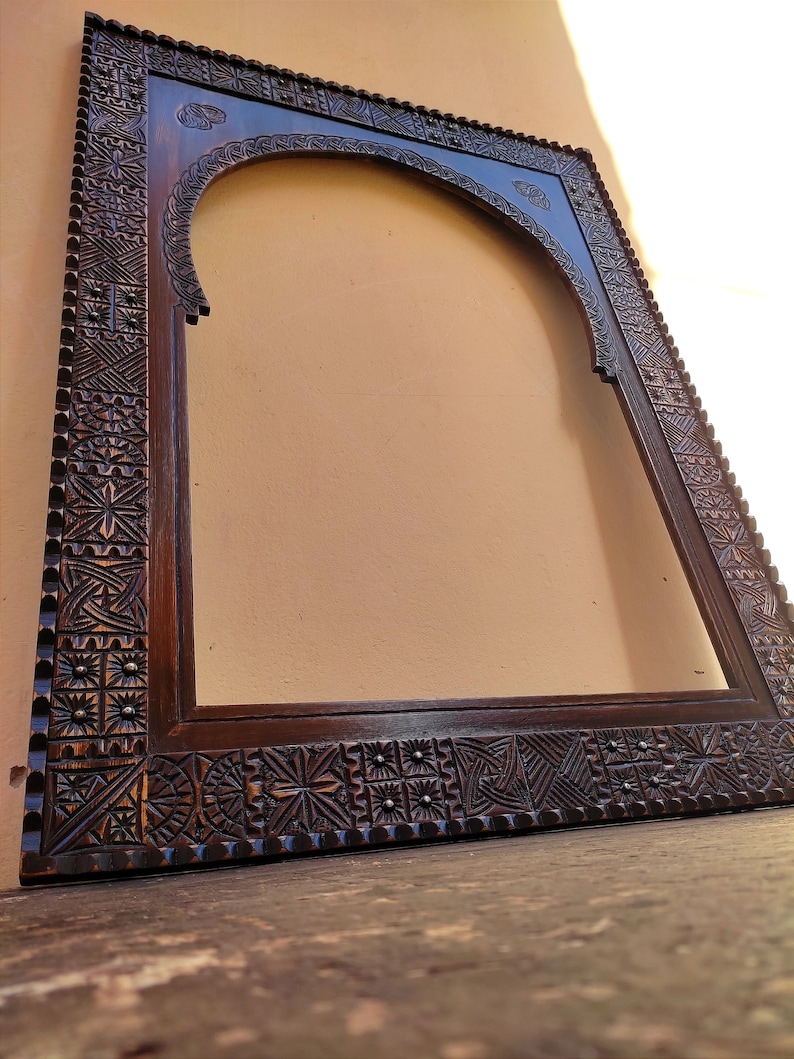May include: Dark brown wooden frame with intricate carved geometric patterns. The frame has an arched top and a rectangular shape, with detailed carvings along the edges. The wood has a rich, warm tone, and the frame appears to be empty.