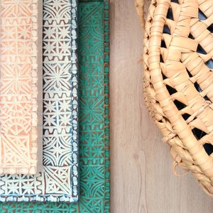 May include: Three decorative wooden trays with intricate geometric patterns. The top tray is peach, the middle tray is white with blue accents, and the bottom tray is green. The trays are stacked on top of each other.