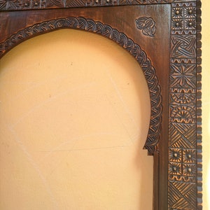 May include: A dark brown, intricately carved wooden frame with an arched top. The frame features detailed geometric and floral patterns, with small brass accents. The design suggests a vintage or antique style, suitable for a mirror or artwork.
