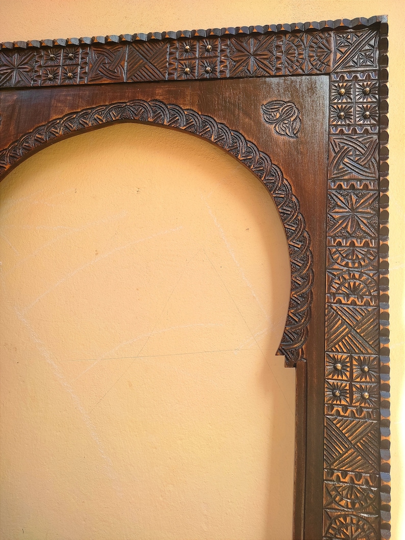 May include: A dark brown, intricately carved wooden frame with an arched top. The frame features detailed geometric and floral patterns, with small brass accents. The design suggests a vintage or antique style, suitable for a mirror or artwork.