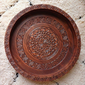 May include: A round, wooden tray with intricate carved geometric patterns. The tray is a warm brown color and features a central starburst design surrounded by concentric circles of triangles and other shapes. The rim also has a carved design.