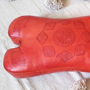 May include: A red leather pouf with intricate embossed geometric patterns. The pouf is shaped like a large, rounded pillow with two rounded ends.