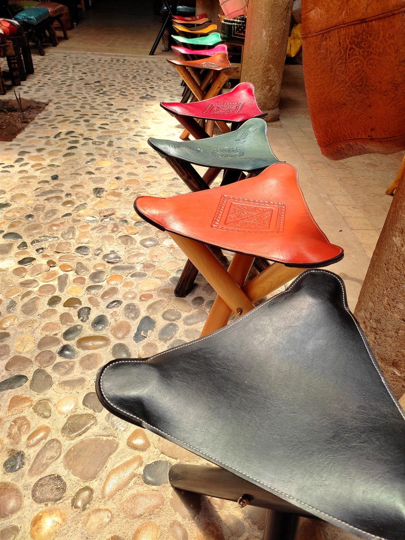 May include: A row of colorful leather Moroccan poufs, or ottomans, with intricate designs. The poufs are arranged in a line on a stone floor. The poufs are made of leather and have a wooden frame.