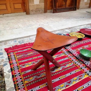 May include: A brown leather folding stool with a decorative stitched design sits on a red and black patterned rug.