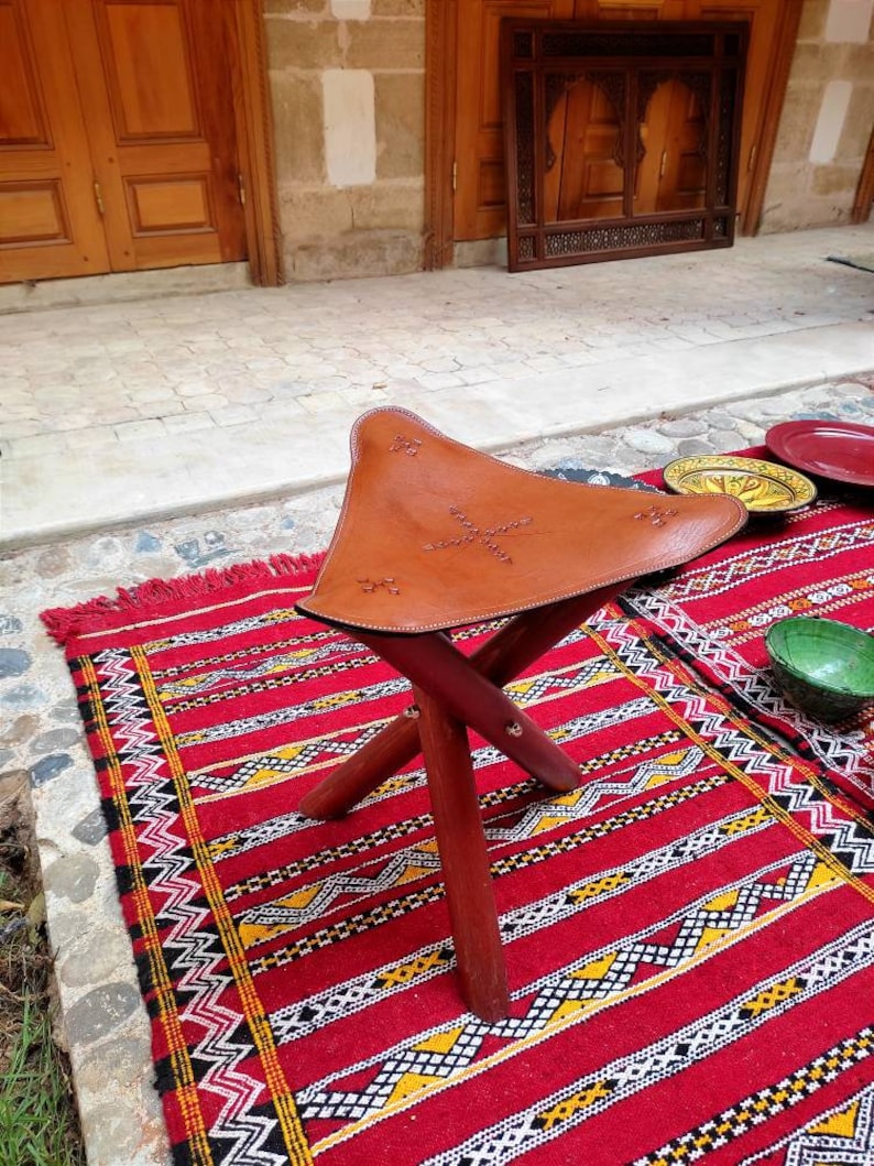 May include: A brown leather folding stool with a decorative stitched design sits on a red and black patterned rug.