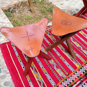 May include: Two brown leather triangular stools with intricate stitching and wooden legs. The stools are on a red and white patterned rug.