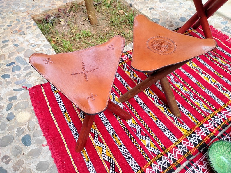 May include: Two brown leather triangular stools with intricate stitching and wooden legs. The stools are on a red and white patterned rug.
