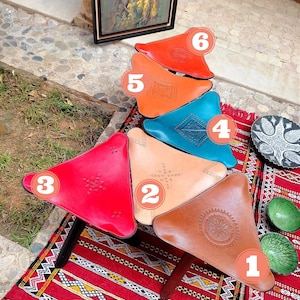 Op de afbeelding: Zes driehoekige leren poefs in verschillende kleuren, waaronder rood, oranje, bruin, beige, blauw en rood. De poefs zijn in piramidevorm gerangschikt op een rood-wit gepatroneerd tapijt. De poefs zijn versierd met ingewikkelde stiksels en ontwerpen. Wat is je favoriete kleur?