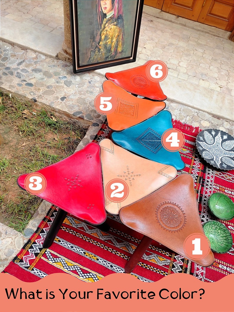 Op de afbeelding: Zes driehoekige leren poefs in verschillende kleuren, waaronder rood, oranje, bruin, beige, blauw en rood. De poefs zijn in piramidevorm gerangschikt op een rood-wit gepatroneerd tapijt. De poefs zijn versierd met ingewikkelde stiksels en ontwerpen. Wat is je favoriete kleur?