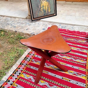 May include: A brown leather three-legged stool with a decorative design on the seat. The stool is sitting on a red and white patterned rug.