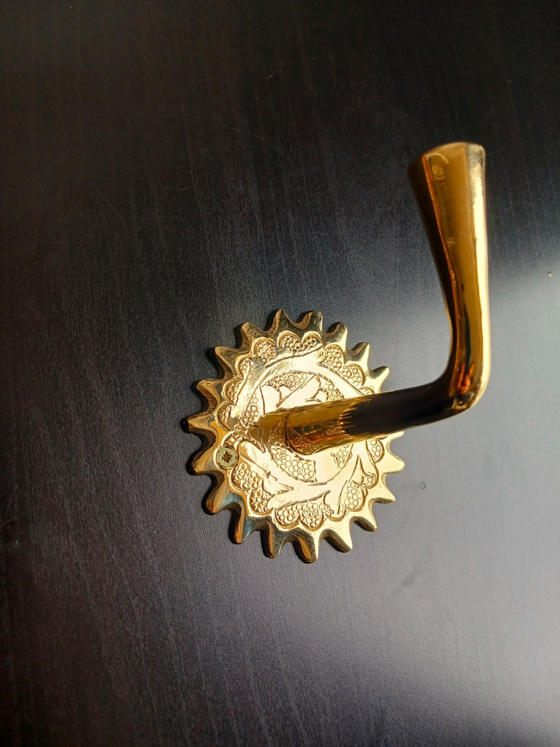 May include: A gold-toned metal hook with a decorative, starburst-shaped base. The hook is attached to a dark surface.