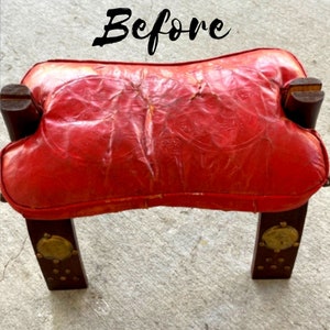 May include: A red leather ottoman with wooden legs and brass accents. The ottoman has a worn, distressed look.
