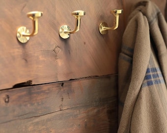 May include: Three gold metal hooks mounted on a wooden wall. A brown and blue plaid scarf is hanging on one of the hooks.
