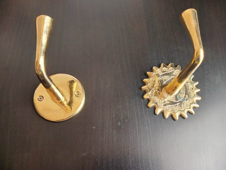 May include: Two gold-toned wall hooks with decorative details. One hook has a round base with a smooth surface. The other hook has a starburst-shaped base with intricate designs.