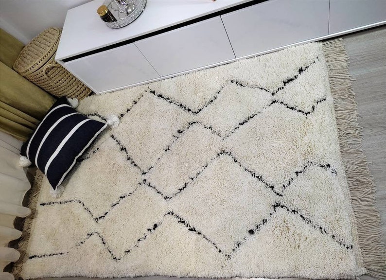 May include: A white shag rug with a black diamond pattern. The rug has a fringe border and is laid out on a hardwood floor.