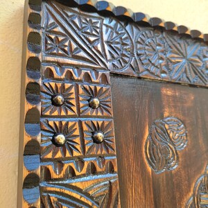 May include: Close-up of a dark brown wooden frame with intricate carved floral and geometric patterns. The frame has a scalloped edge and is accented with small, round metal studs. The wood grain is visible, adding to the rustic aesthetic.