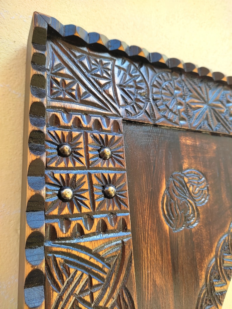 May include: Close-up of a dark brown wooden frame with intricate carved floral and geometric patterns. The frame has a scalloped edge and is accented with small, round metal studs. The wood grain is visible, adding to the rustic aesthetic.